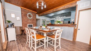 A dining area with a wooden table and chairs, a brick wall, and a kitchen in the background.