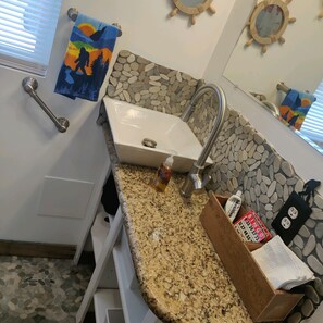 A bathroom with a granite countertop, a white sink, and a stone backsplash. There is a towel rack with a colorful towel, a wooden box with toiletries, and a mirror with a wooden frame.