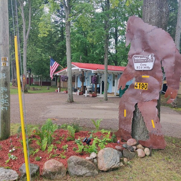 A rustic cabin with a red roof, surrounded by trees and a small garden with rocks and plants. A large welcome sign with the number 4190 is visible.