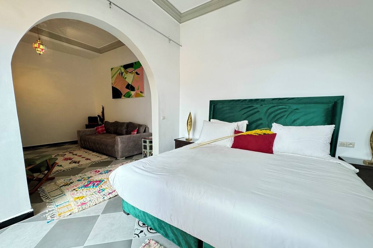 A hotel room with a large bed, a green upholstered headboard, a colorful rug, and a small seating area with a sofa and a side table.