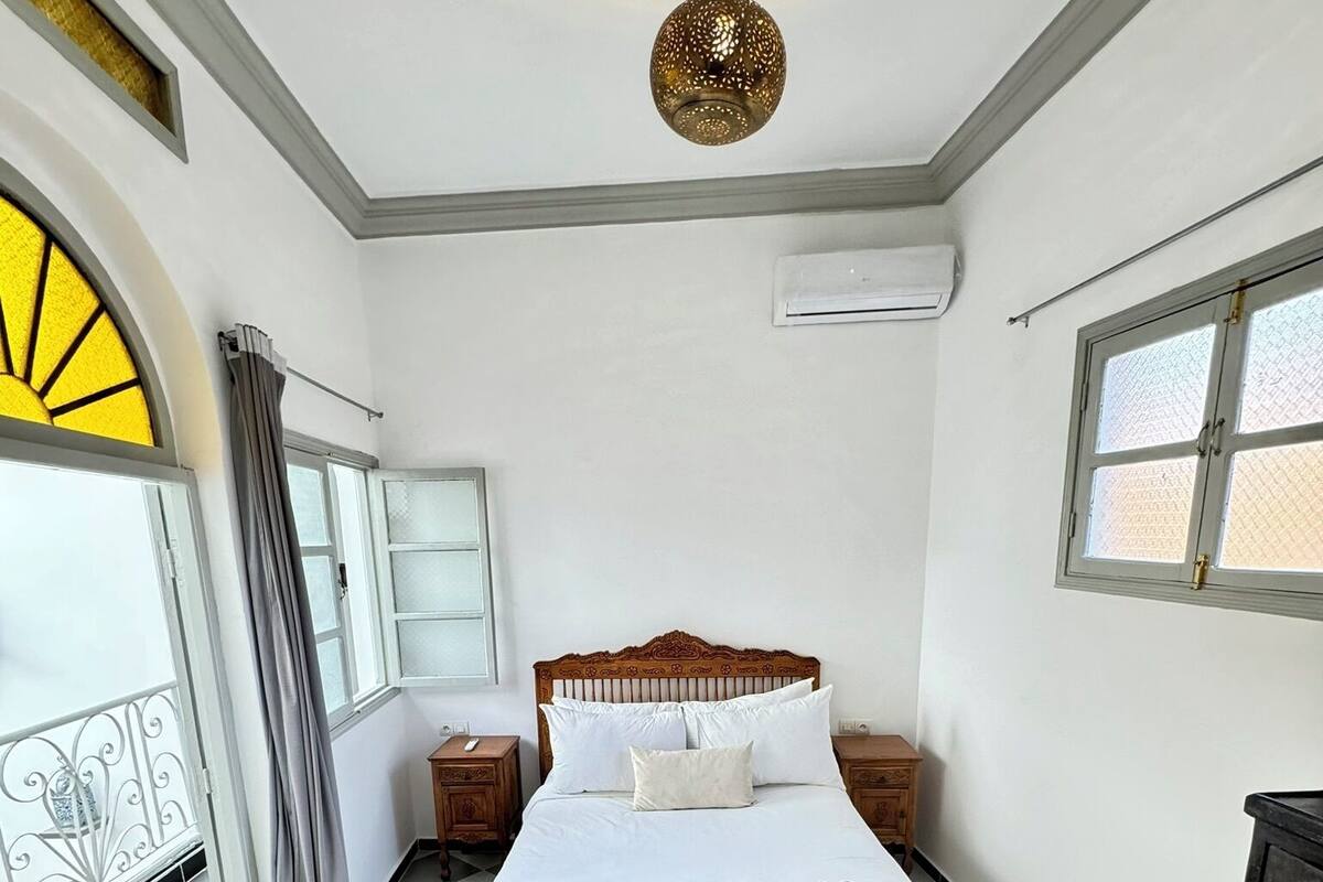 A bedroom with a double bed, a hanging light fixture, a window with a view, and a checkered floor.