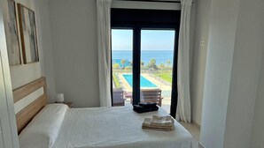 A hotel room with a bed, a view of a pool and beach, and a window with curtains.