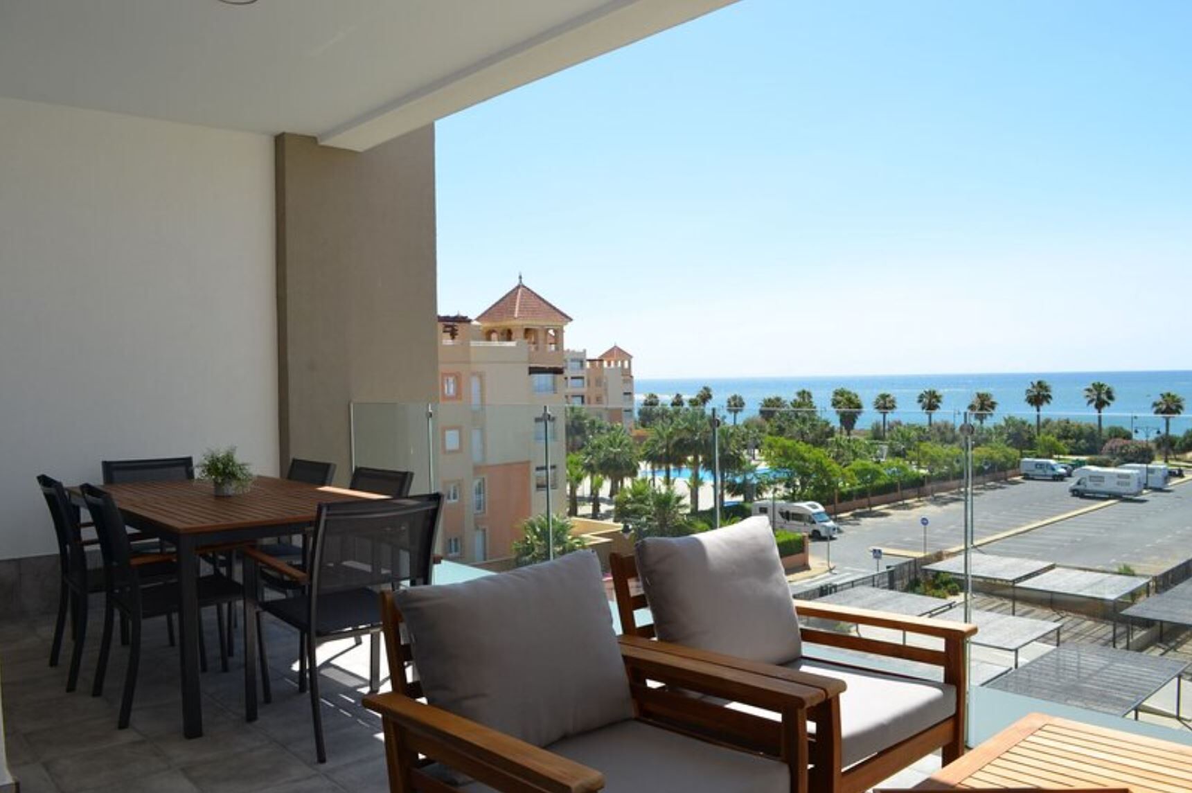 A balcony with a wooden table and chairs, overlooking a beach and a parking area with several vehicles.