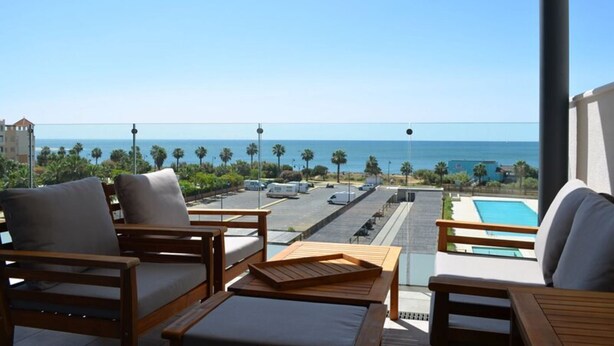A balcony with wooden furniture, a pool, and a view of the ocean and parking area.