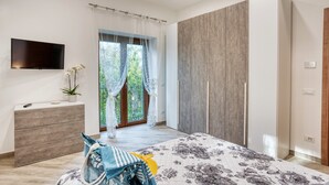 A bedroom with a bed, a TV, a wardrobe, a bedside table with a plant, and a window with curtains. - Margaret House Sorrento (Sorrento)
