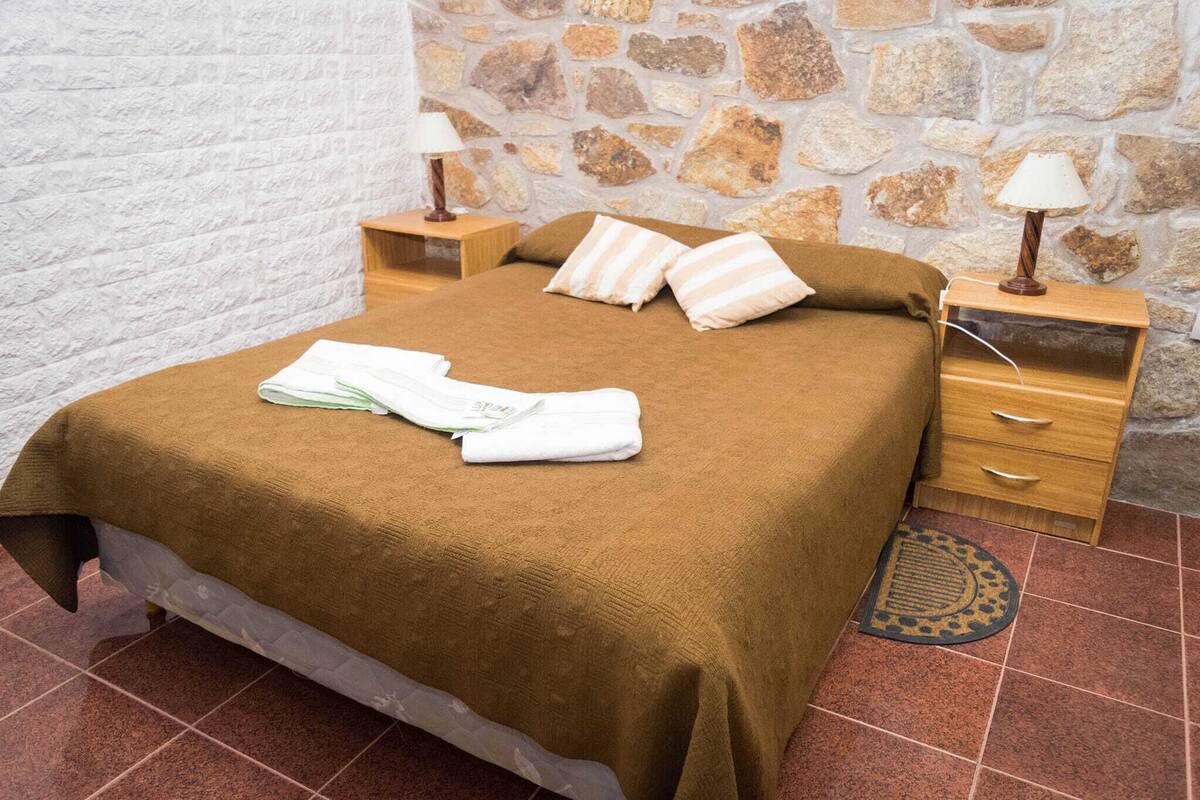 A neatly made bed with a brown blanket, two pillows, and a folded towel on a wooden nightstand with a lamp. The room has a stone wall and tiled floor.