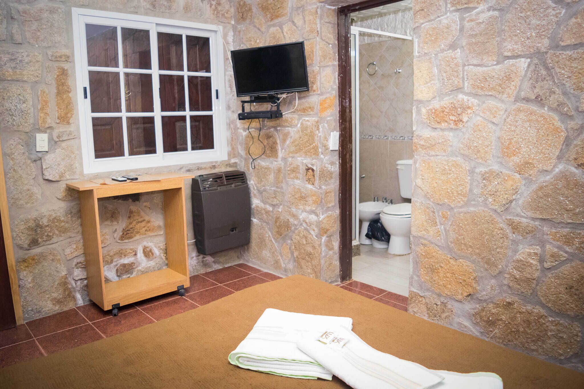 Un baño con pared de piedra, tocador de madera, televisor montado en la pared y un inodoro visible a través de una puerta de cristal.