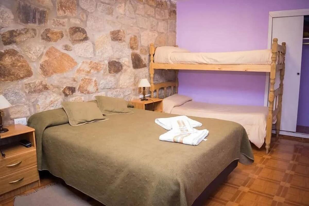 A room with a bunk bed, a single bed, a stone wall, a wooden nightstand, and a bedside table with a lamp.