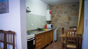 A compact kitchen with a gas stove, microwave, and a small refrigerator. There is a wooden dining table with chairs and a stone wall in the background.