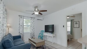 A living room with a blue sofa, a coffee table, a TV on a white stand, patterned curtains, and a ceiling fan. - Downtown Dunedin Cottage • Walk to Restaurants (Dunedin)
