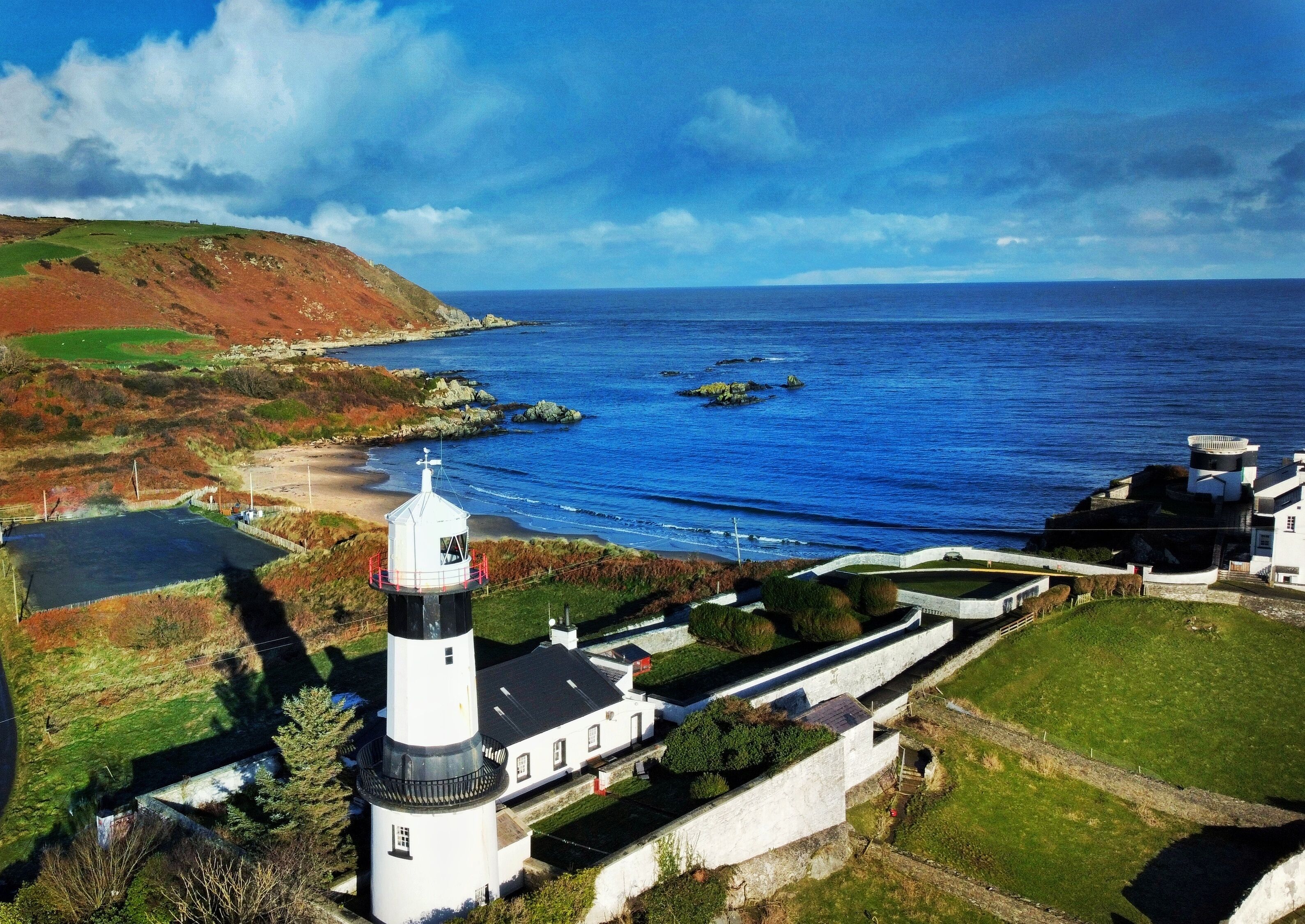 Un faro con una torre bianca e nera, posto su una collina erbosa che si affaccia su una zona costiera con mare cristallino e affioramenti rocciosi.