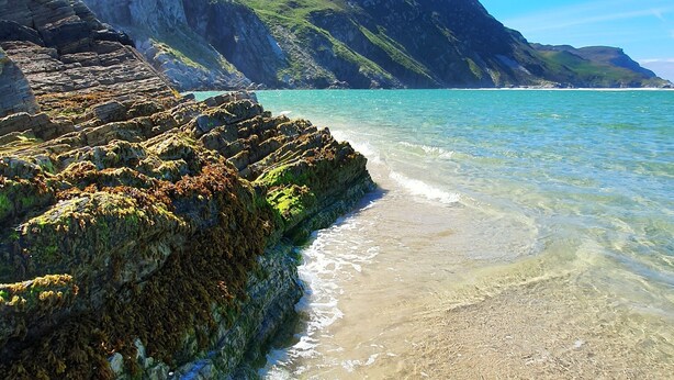 Ein Küstenstrand mit klarem Wasser, steinigen Klippen und einem gebirgigen Hintergrund.