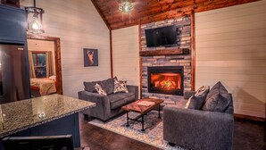 A cozy living room with a stone fireplace, a television mounted above it, and a kitchen area with a granite countertop and bar stools. - Romantic Couples Cabin minutes from Casino Excitement & Lakeside Serenity (Broken Bow)