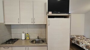 Een compacte kitchenette met een fornuis, spoelbak en kasten, een flatscreen-tv bevestigd boven de kasten, en een bed met een bloemenbedekking in de aangrenzende kamer.