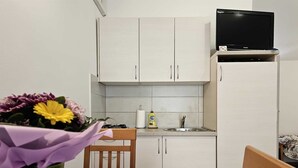 Een kleine keuken met witte kastjes, een gasfornuis, een spoelbak en een zwarte televisie bovenop een witte koelkast.
