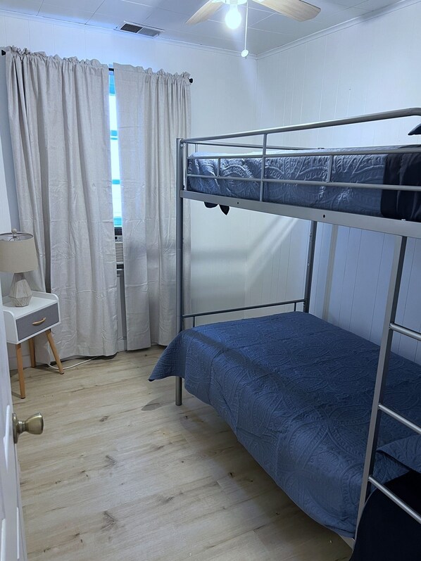 Habitació amb un llit búnquer, una finestra, cortines, una tauleta de nit i un ventilador de sostre.