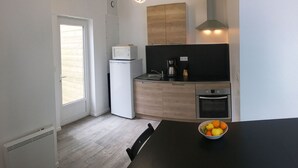 Eine kompakte Küche mit Kühlschrank, Herd und Spüle. Ein Fenster mit Jalousien, eine Schale mit Obst auf dem Tisch und ein Stuhl.