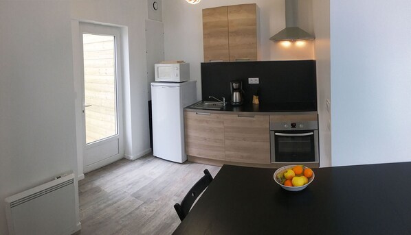 A compact kitchen with a refrigerator, stove, and sink. There's a window with blinds, a bowl of fruit on the table, and a chair.