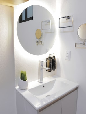 A modern bathroom with a white sink, a round mirror, and two wall-mounted soap dispensers.