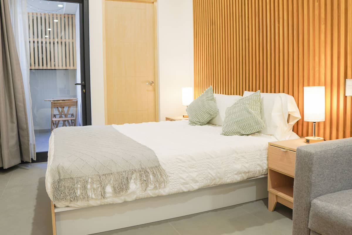 A bedroom with a bed, bedside tables, a chair, a wooden door, and a view of a balcony.