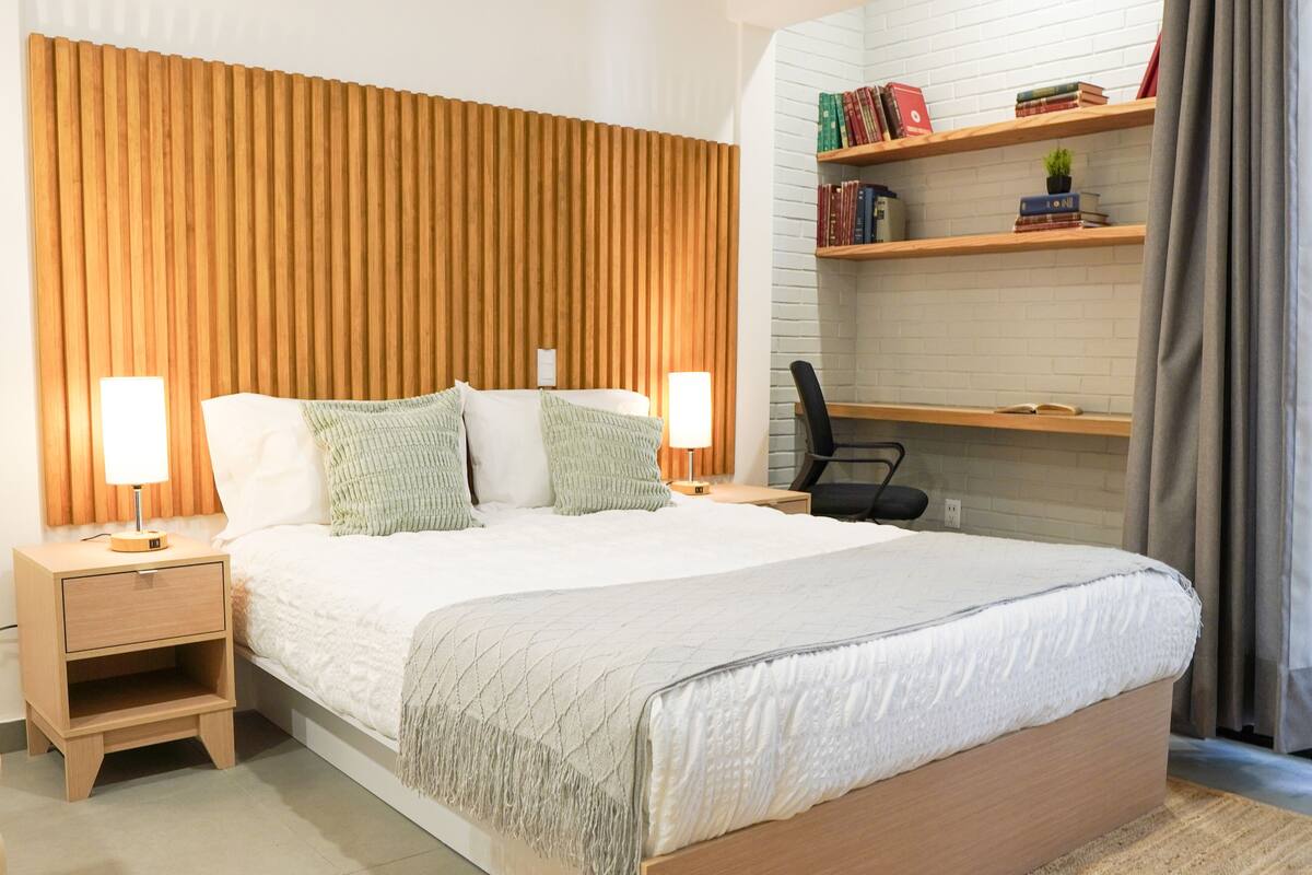 A neatly arranged bedroom with a wooden headboard, a bed with white bedding, two bedside tables with lamps, a small shelf with books, and a chair.