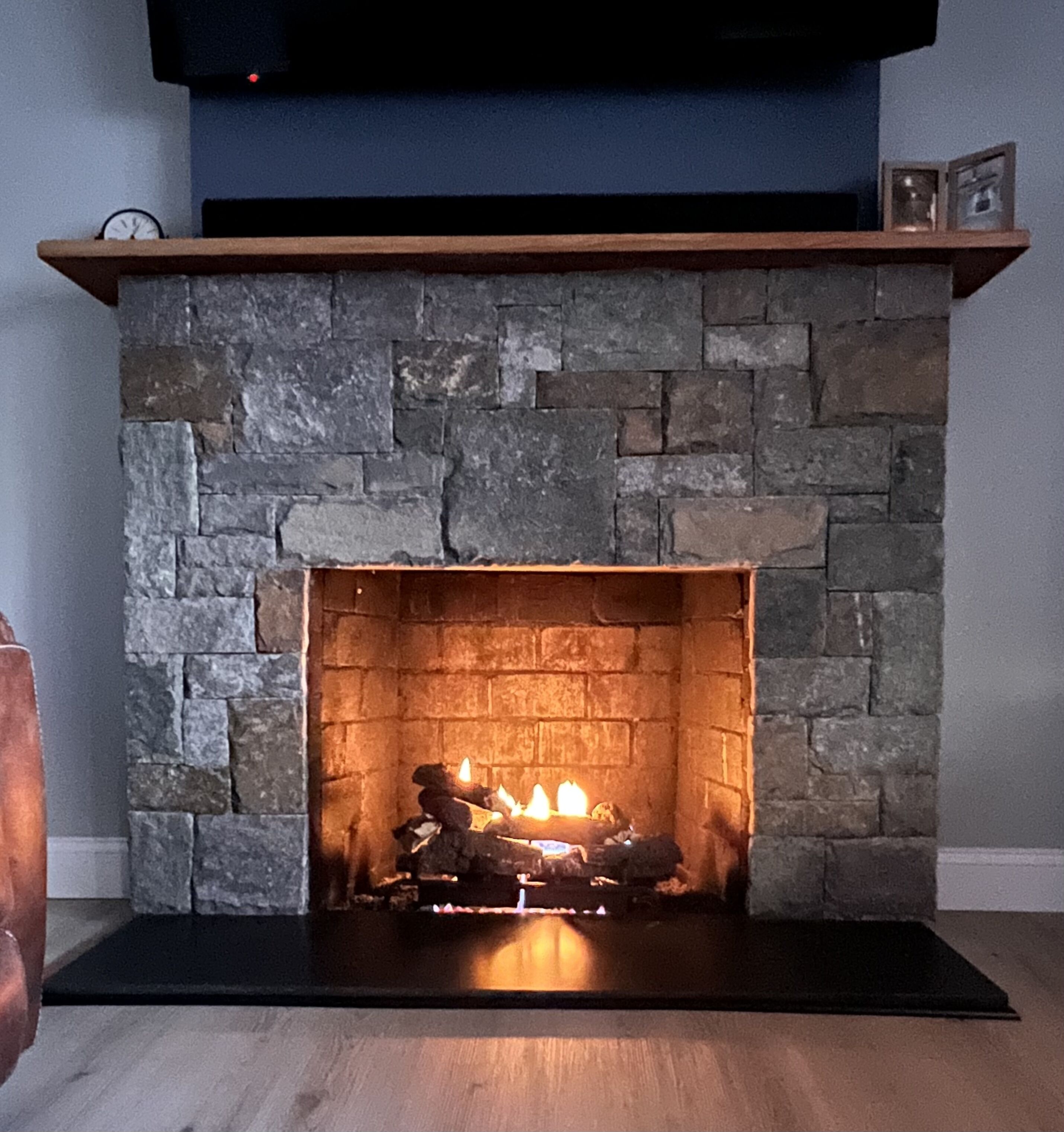 Una chimenea de piedra con repisa de madera, un televisor montado encima y un cuadro enmarcado sobre la repisa.