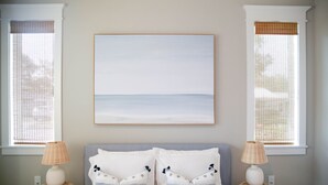 Une chambre à coucher avec un lit, deux tables de chevet, un tableau encadré et deux fenêtres équipées de stores.