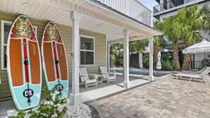 A covered outdoor area with two surfboards leaning against a house, a pool, and lounge chairs.
