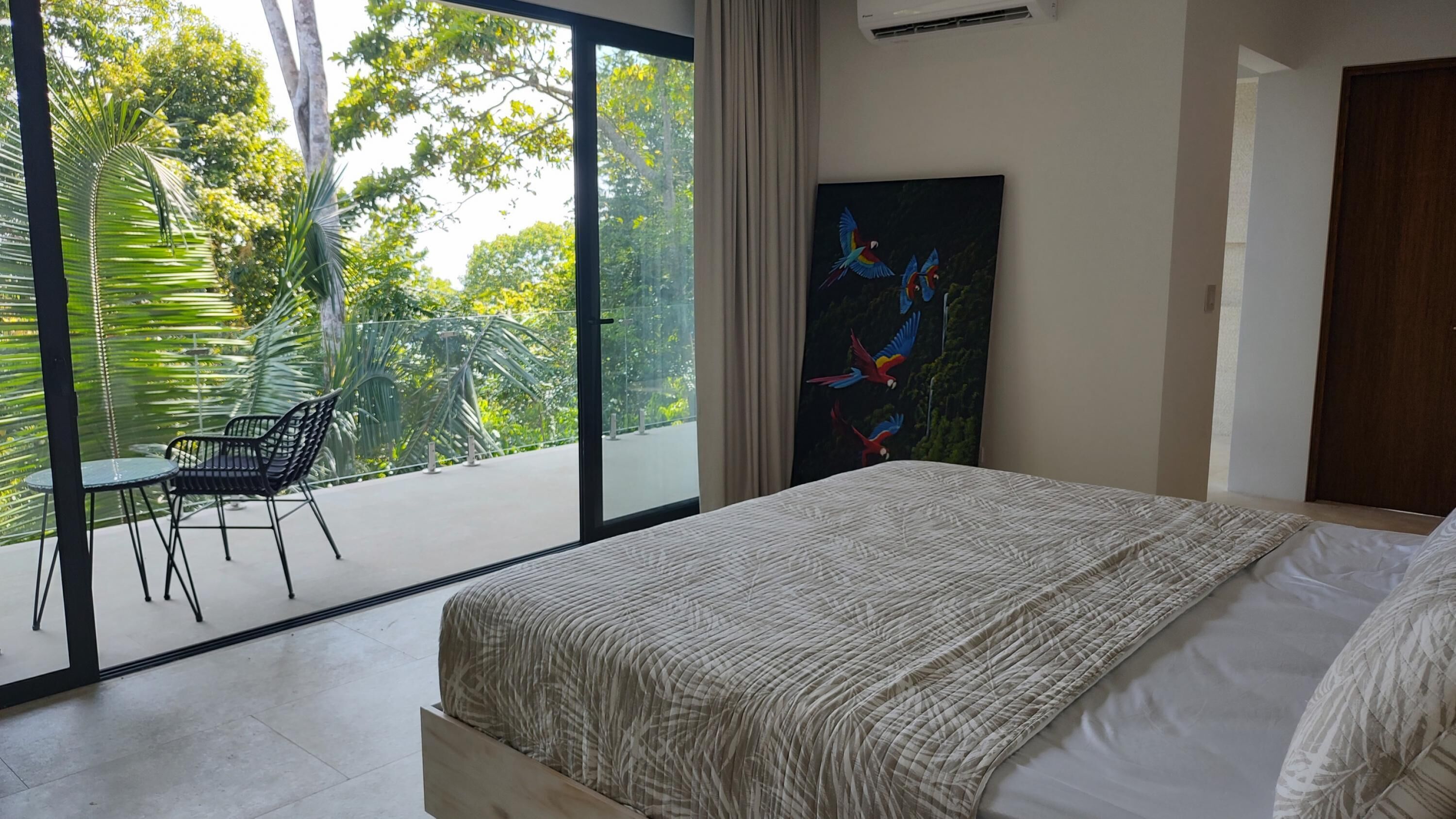 A bedroom with a large bed, a view of greenery through a sliding door, a painting of birds, and a small table with chairs.