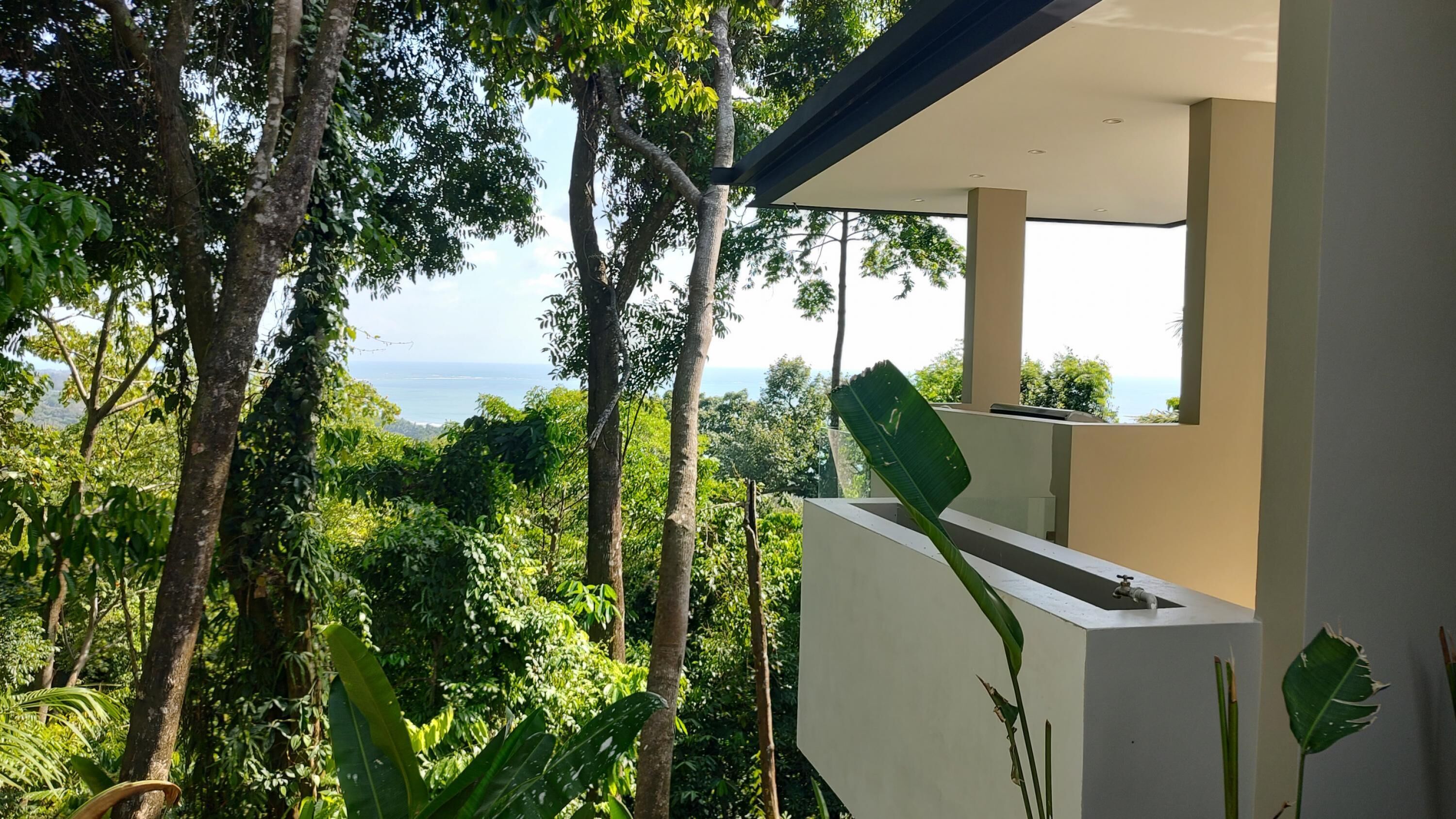 A modern building with a balcony overlooking a lush, green landscape and a distant view of the sea.