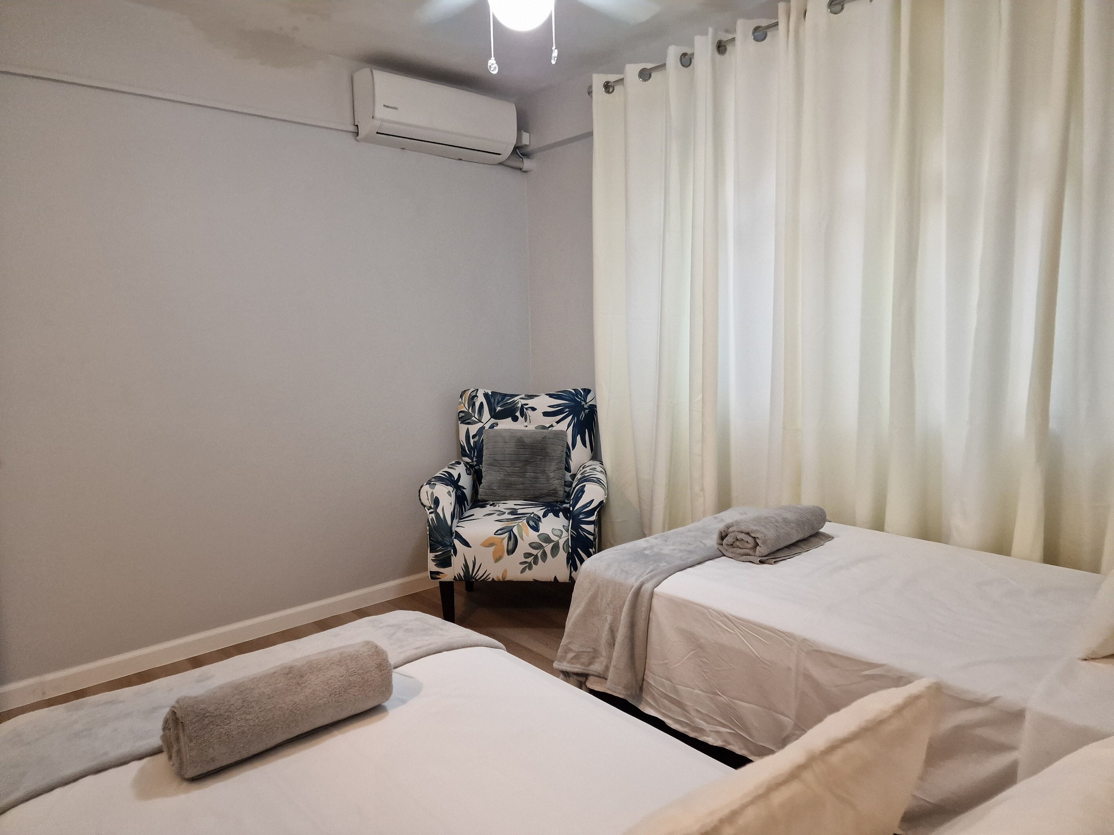A hotel room with two beds, a chair with a floral pattern, a towel on one bed, and a curtain on the window.