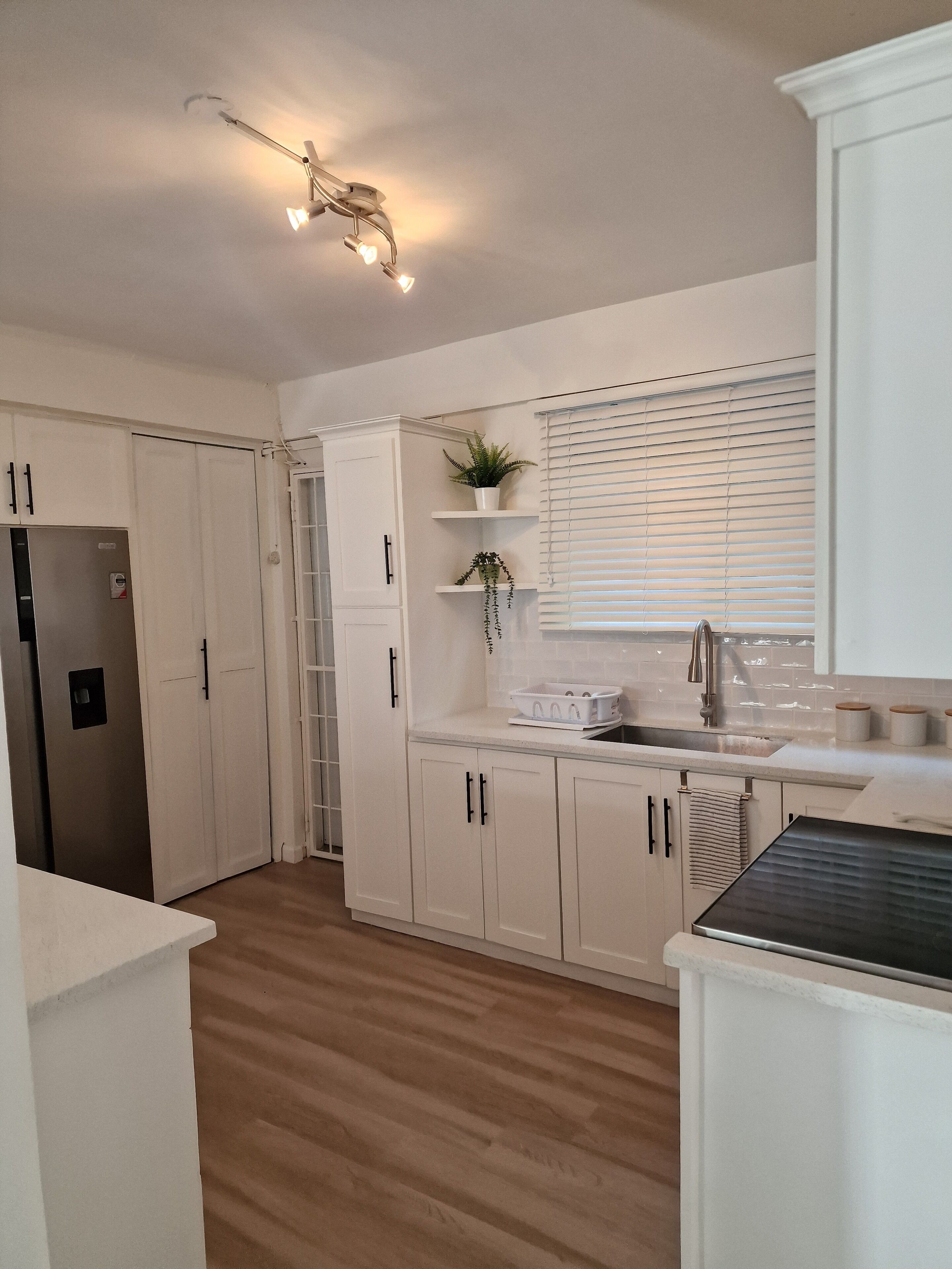 Una cocina moderna con armarios blancos, un refrigerador de acero inoxidable, un fregadero y una ventana con persianas.