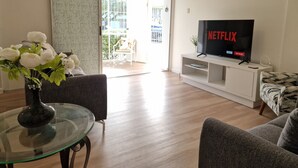 Un salon avec une table basse en verre, deux canapés, un téléviseur affichant le logo « NETFLIX » et une porte-fenêtre coulissante donnant sur un espace extérieur.