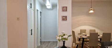 A dining area with a glass table, chairs, and a vase of flowers. There is a hallway leading to another room.