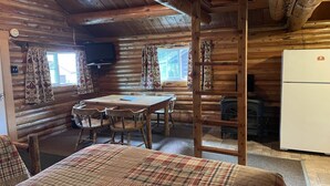 Interno di una baita di legno con zona pranzo, un letto, una scala, un frigorifero e un camino.