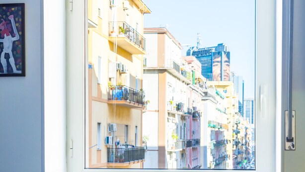 Una mesa con una cafetera, un tarro, naranjas y una taza, con vistas a edificios a través de una ventana.