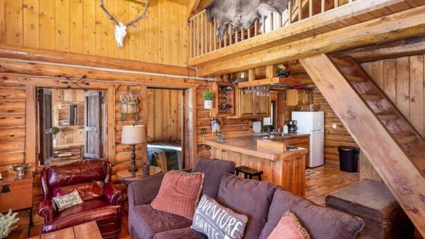 Een rustiek cabinteinterieur met een houten eethoek, een leren bank en een trap naar een hoger niveau.