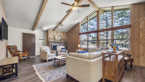 A spacious living room with a high ceiling, wooden beams, a large window with a view of trees, a fireplace, and a cozy seating area. - Charming Home in Country Club Flagstaff with Views (Flagstaff)
