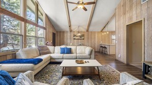A spacious living room with a large window, a ceiling fan, a sofa, a coffee table, and a patterned rug. - Charming Home in Country Club Flagstaff with Views (Flagstaff)