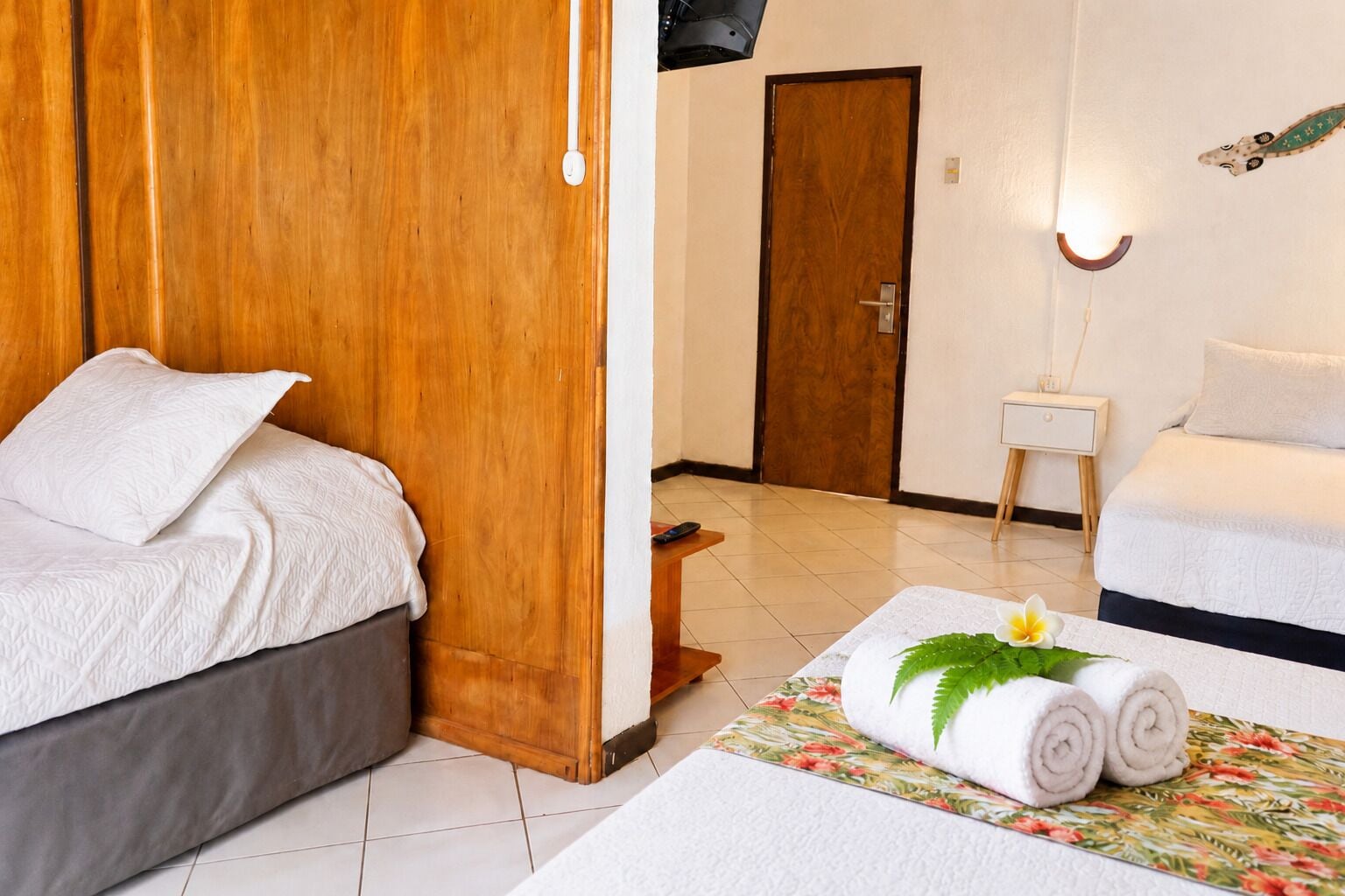 Habitación de hotel con armario de madera, cama con ropa blanca, mesita de noche con lámpara, sofá con cojines blancos y alfombra estampada con motivos florales.