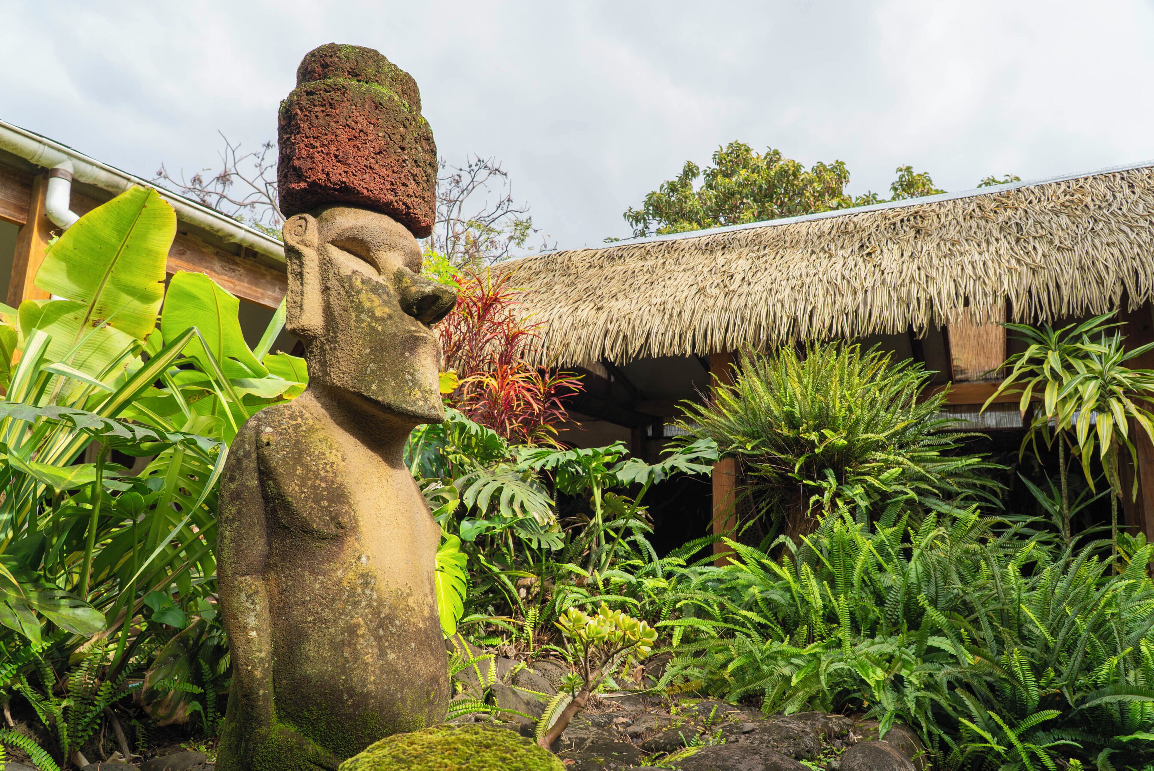 Una estatua de piedra en un jardín exuberante, con plantas tropicales y una estructura de techo de paja.