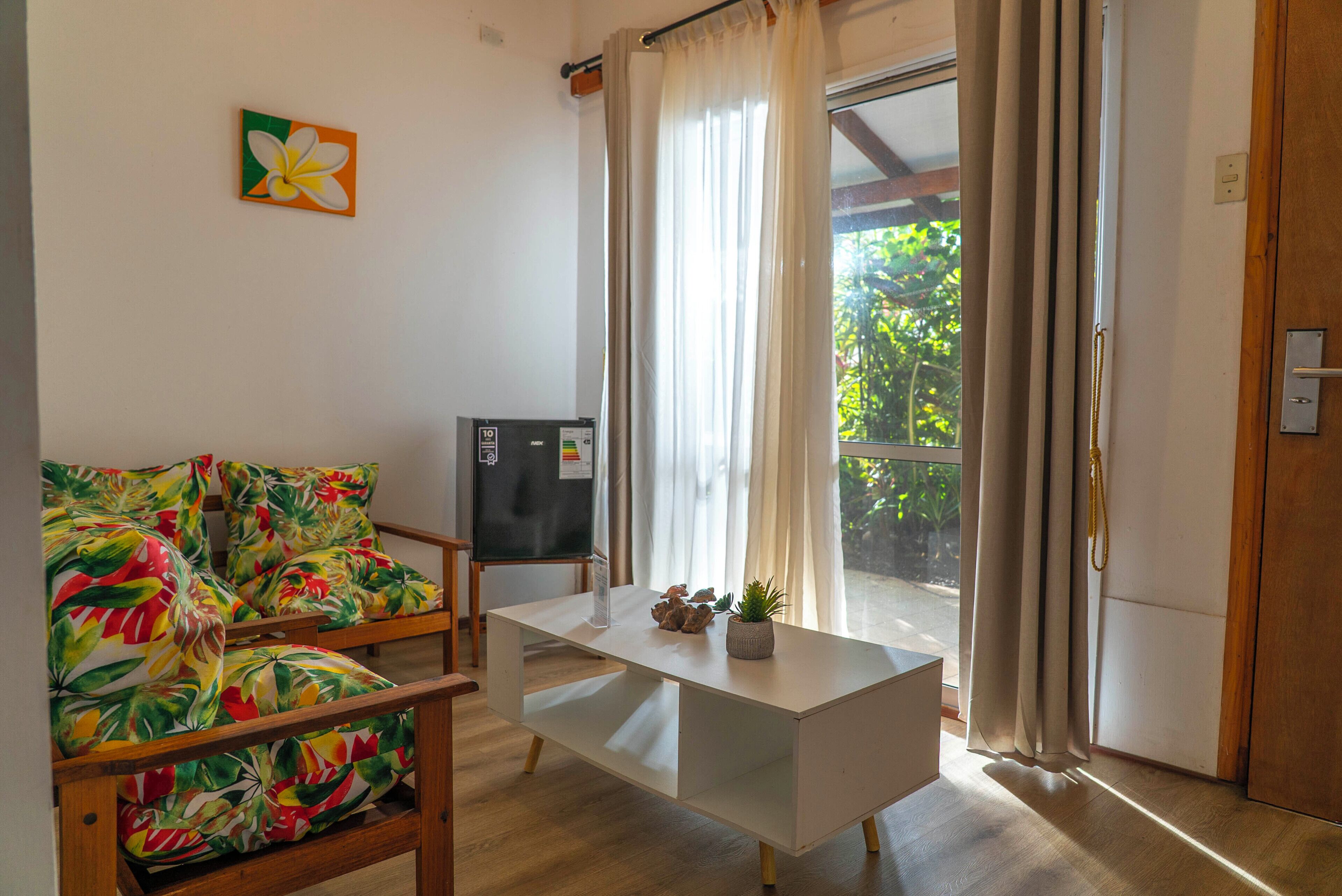 A room with a wooden sofa, a small table, a refrigerator, and a sliding glass door leading to a balcony.