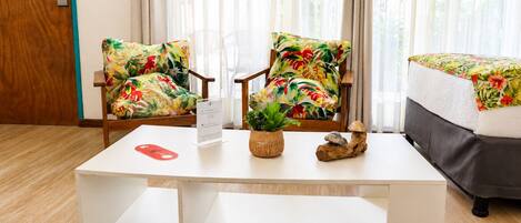 Quarto de hotel com uma mesa de centro branca, duas poltronas com padrão floral, uma cama com colcha floral combinando e vista para o verde através de cortinas translúcidas.
