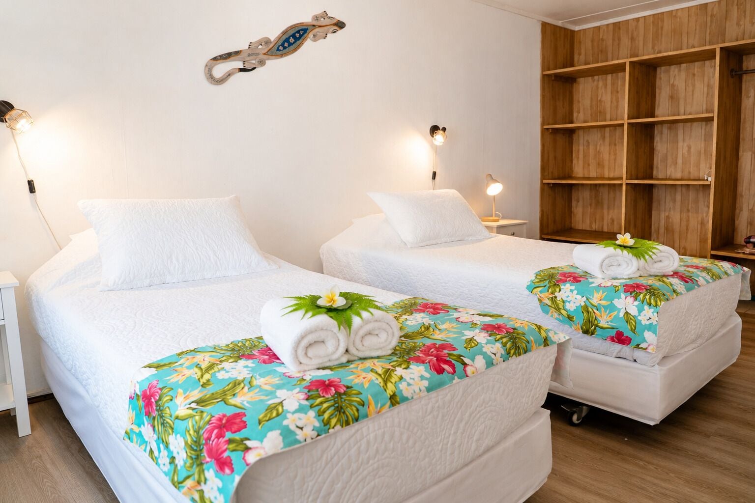 A hotel room with two beds, each with a floral bedspread and rolled towels. There are wall-mounted lamps and a wooden shelf unit.