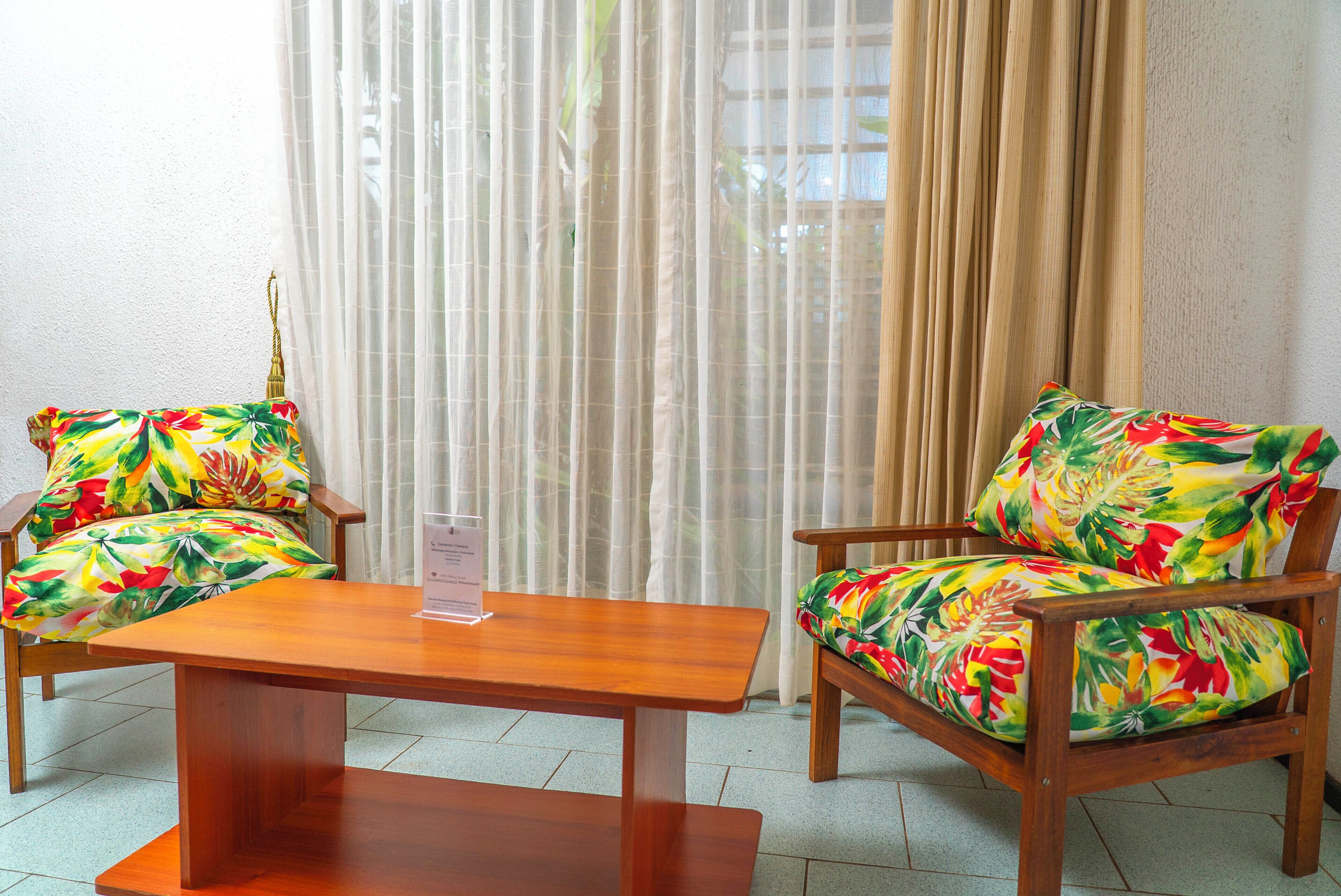 A room with two wooden chairs with floral upholstery, a wooden table, and a window with sheer curtains.