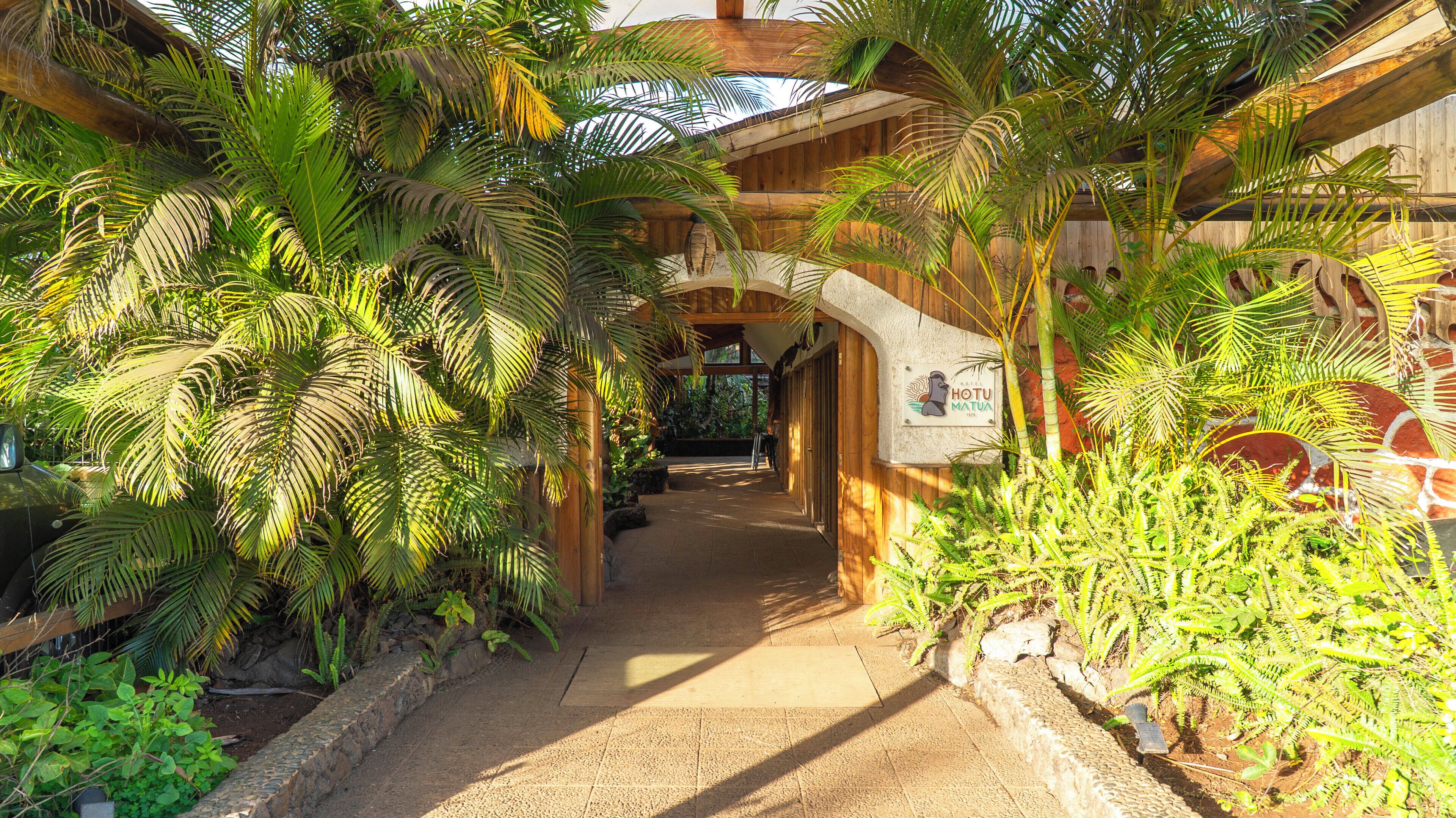 An indoor pathway with wooden beams and tropical plants, leading to a wooden structure with a sign.