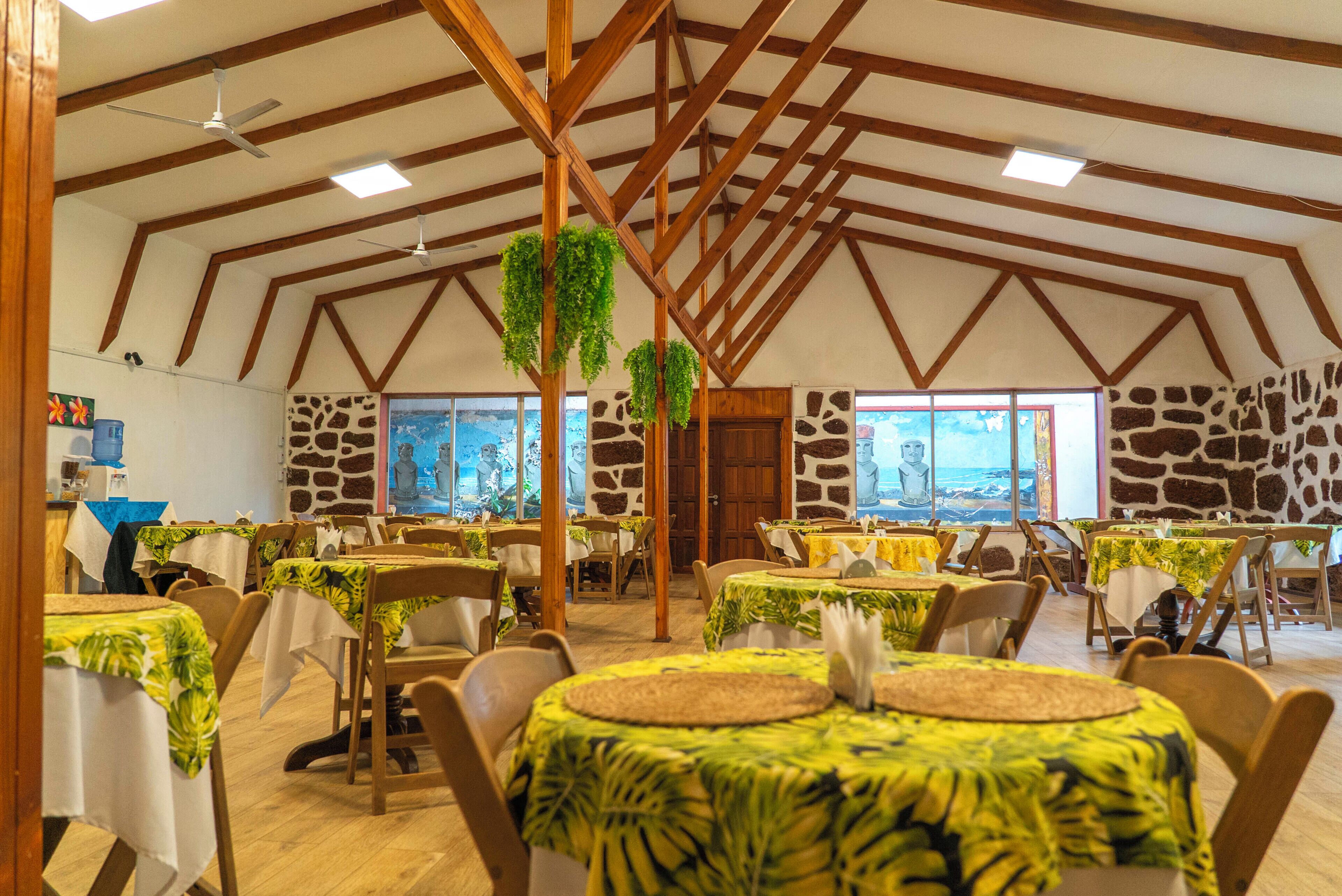 Un restaurante con altos vigueros de madera, paredes de piedra y mesas cubiertas con manteles estampados de hojas tropicales.