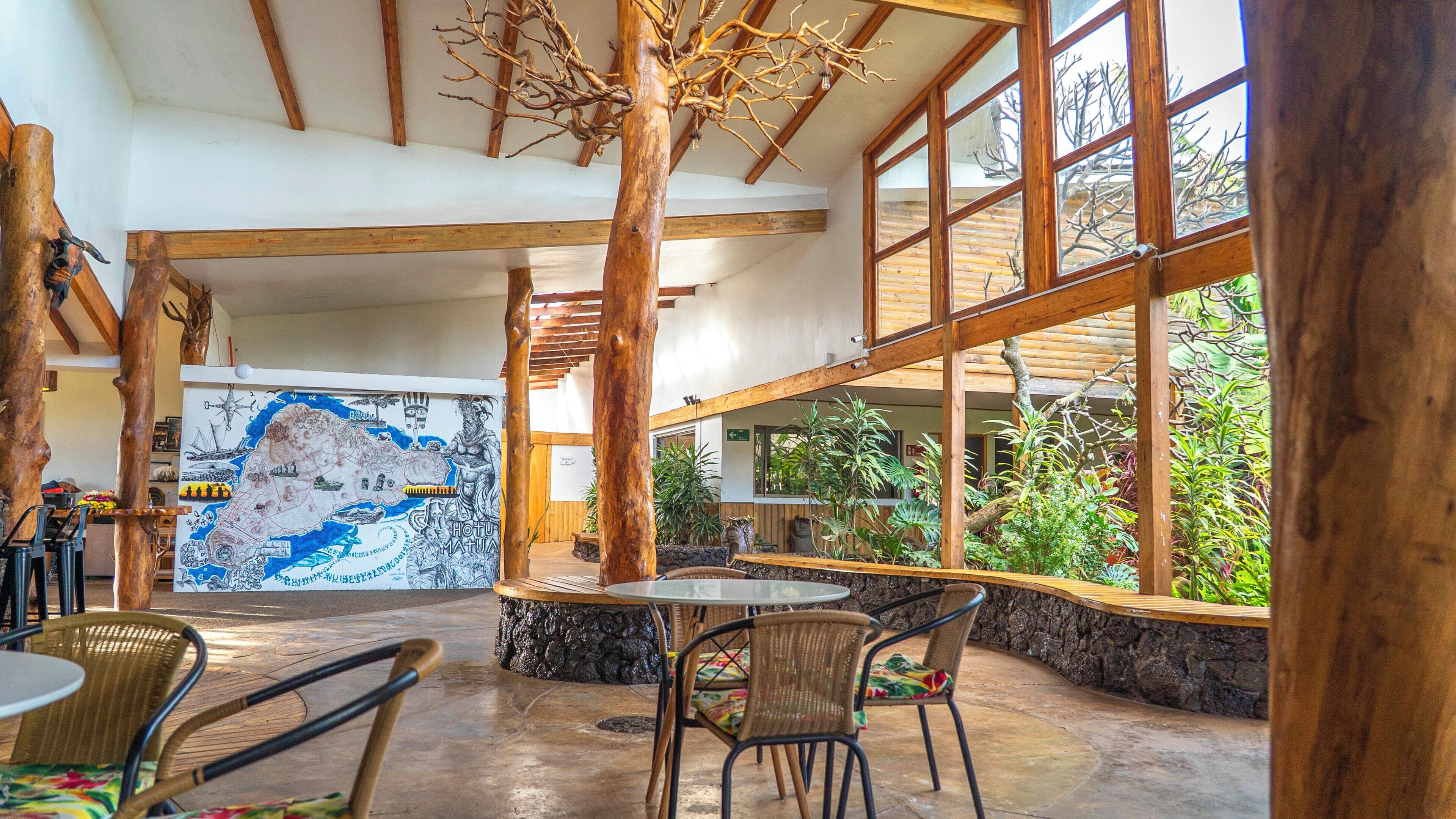 A spacious indoor area with a high ceiling, wooden beams, and large windows. There is a mural on the wall and a seating area with tables and chairs.