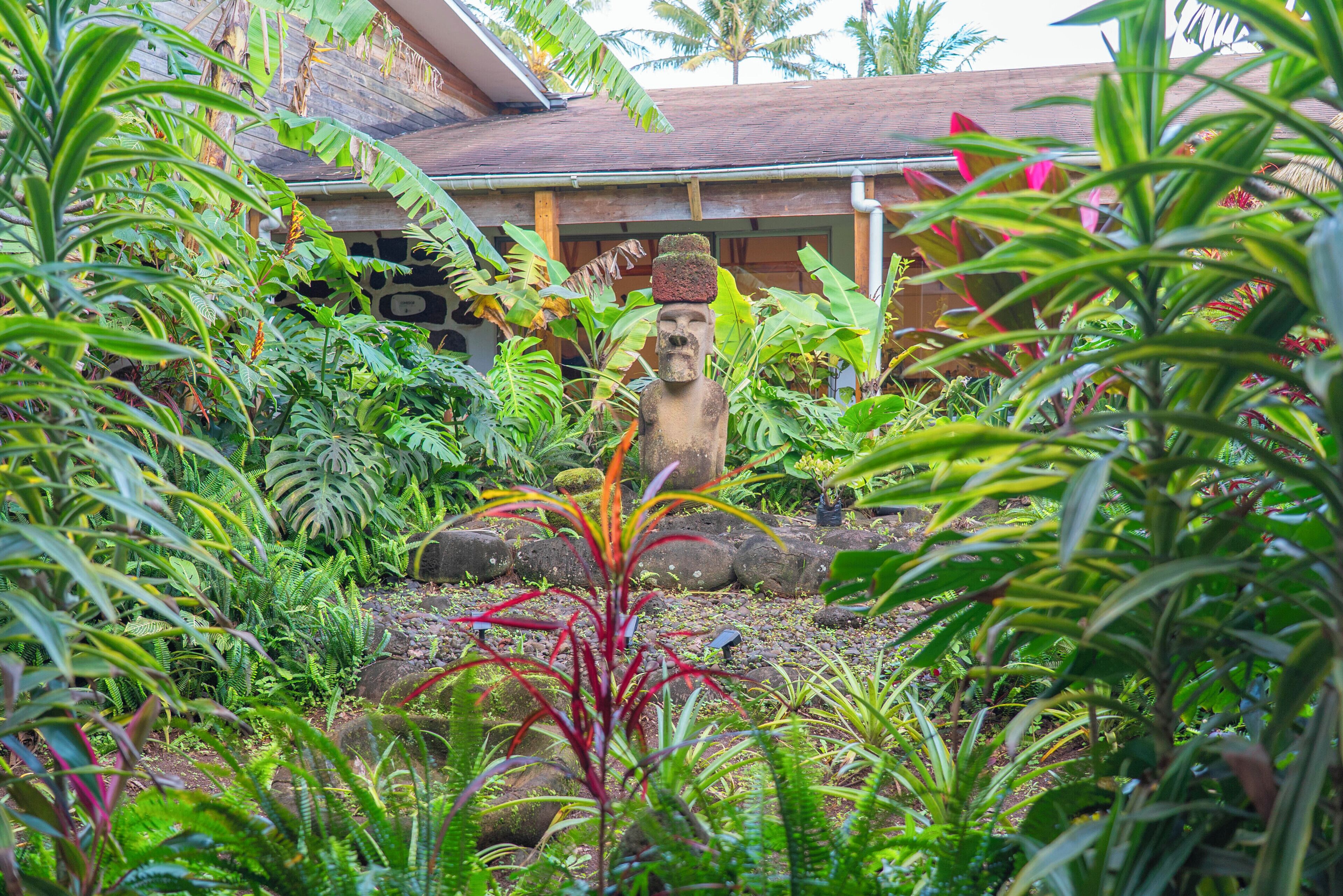 Un jardín con una estatua de piedra, abundante vegetación y un edificio de techo inclinado al fondo.