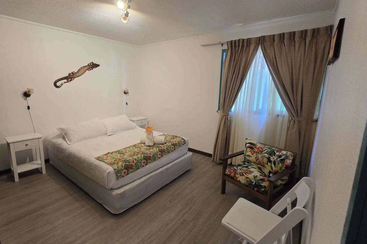A bedroom with a bed, a chair, a nightstand, a wall-mounted lamp, and a window with curtains.